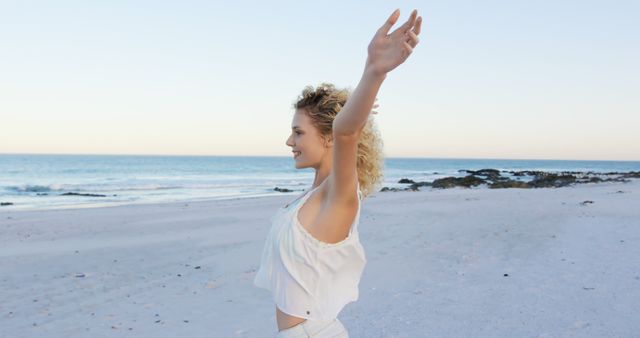 Woman with curly hair rejoicing with outstretched arms on a serene beach during dusk. Capturing a moment of pure joy and relaxation with the ocean waves in the background. Ideal for themes of freedom, happiness, relaxation, escape, and nature's beauty. Great for use in wellness, travel, lifestyle, and motivational content.