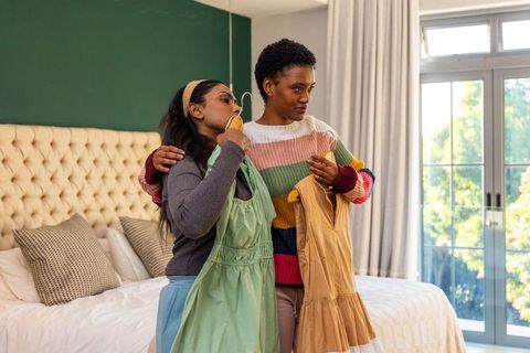 Diverse friends comparing dresses in cozy bedroom with tufted headboard and natural light