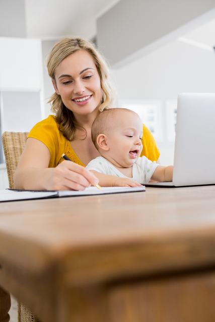Mother writing on a diary while baby boy playing with laptop in kitchen at home