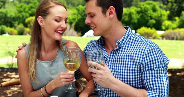 A young Caucasian couple enjoys a romantic moment with glasses of wine in an outdoor setting, with copy space. Their cheerful interaction suggests a celebration or a casual, intimate date in a natural environment.