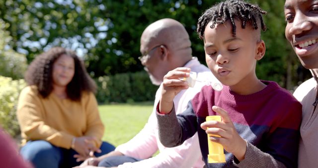 Family members enjoying outdoor playtime in a sunny park. Boy blowing bubbles while others watch with smiles. Great for concepts of togetherness, childhood joy, leisure time, family bonding, and summer activities.