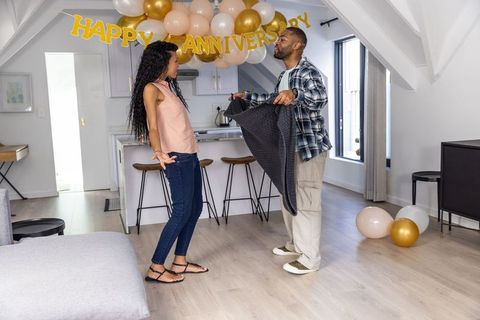 Couple celebrating anniversary with balloons and man offering cozy blanket in modern loft