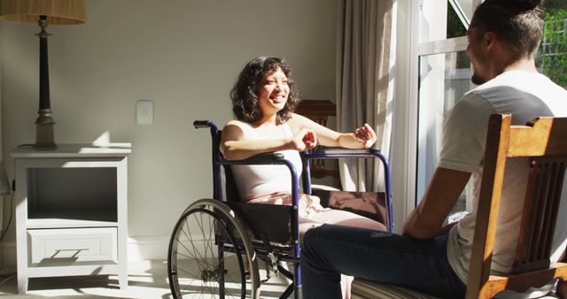 Biracial woman in wheelchair and male partner sitting and talking by window in sunny room. wellbeing and domestic lifestyle with physical disability.