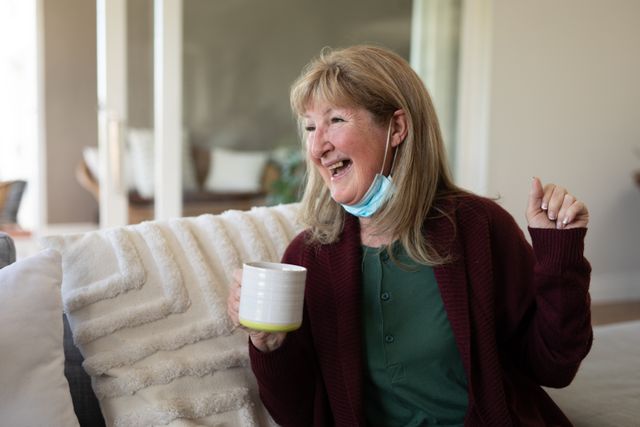 Senior Caucasian woman sitting on a couch, holding a mug, and smiling with a face mask on her chin. Ideal for use in articles or advertisements related to social distancing, Covid-19 safety measures, elderly care, home comfort, and pandemic lifestyle.