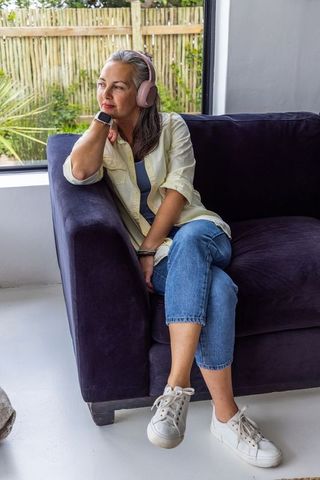 Mature woman relaxing on sofa wearing pink headphones and smartwatch, gazing out window
