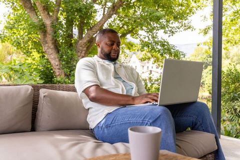 African american man working on laptop on porch sofa with coffee in garden sunlight