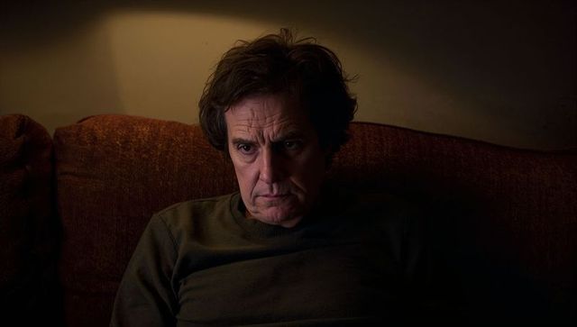 Middle-aged man sitting on rust sofa gazing downward in dim living room, pensive mood