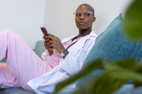African american nurse relaxing on sofa in pink scrubs using smartphone
