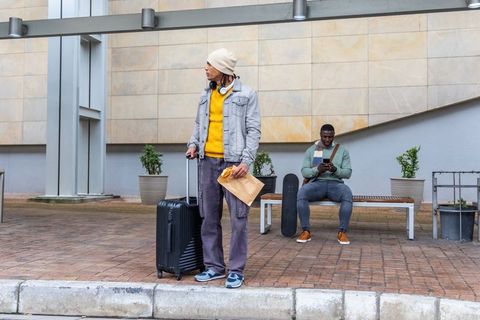 Diverse commuters waiting at urban curb with suitcase, skateboard and smartphone