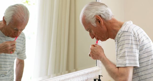 Elderly man brushing his teeth while looking in the bathroom mirror. Useful for promoting senior health, personal hygiene products, or daily routines. Perfect for healthcare, lifestyle, and senior-focused advertisements.
