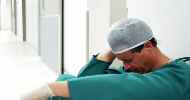 Image depicting an exhausted surgeon sitting down in a hospital hallway, wearing scrubs, surgical cap, and gloves. Suitable for conveying themes of healthcare worker burnout, stress, fatigue, and emotional strain. Ideal for use in articles on medical profession challenges, healthcare worker welfare, and the impacts of long working hours.