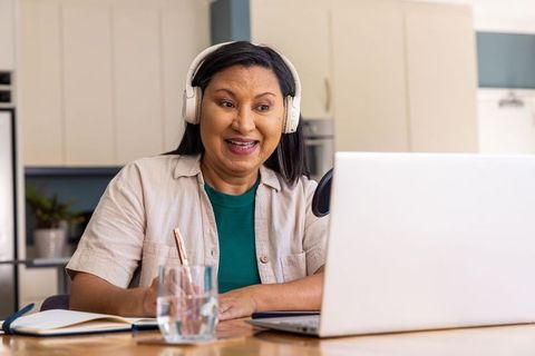 Mature woman hosting podcast from home kitchen wearing headphones and speaking into laptop