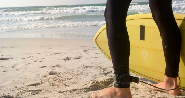 A surfer, barefoot with wetsuit, standing on beach, holding yellow surfboard, leash attached to ankle, waves in background. Ideal for content related to surfing, beach activities, adventure sports, vacation spots, seaside leisure, outdoor hobbies, and fitness tourism.
