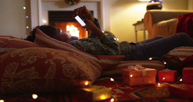 Child lying on cushions using smartphone beside cozy fireplace. Presents and decorative lights create festive atmosphere suggesting relaxing holiday moments at home. Useful for themes of home celebration, technology, and festive relaxation.