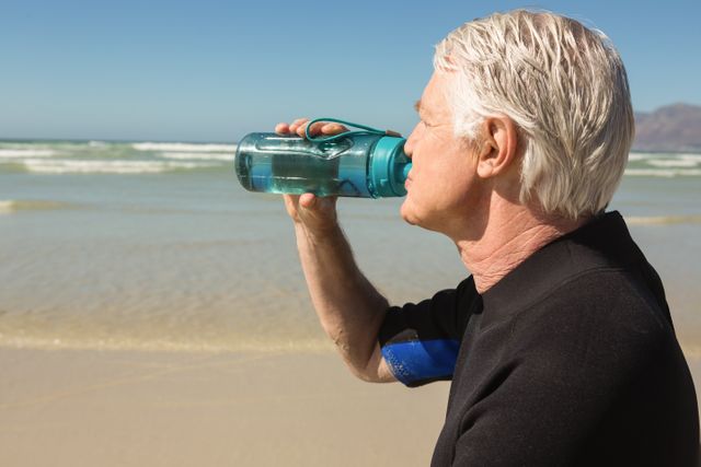 Senior man drinking water from a bottle while at the beach. Ideal for promoting healthy lifestyle, hydration, outdoor activities, and wellness for elderly individuals. Can be used in advertisements for health products, retirement living, and summer activities.