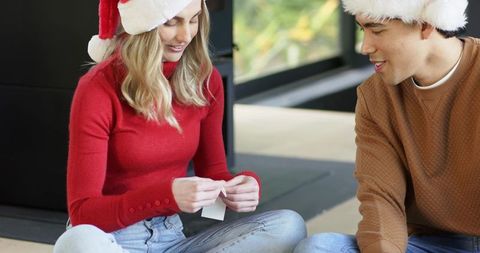 Couple wearing santa hats sitting on floor exchanging holiday note by window with greenery