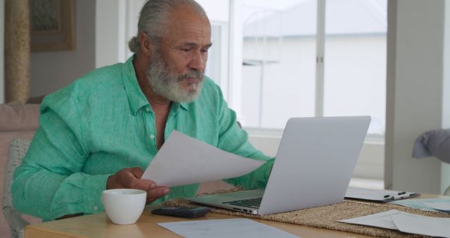 Senior biracial man reviews documents at home office. He's focused on work with a laptop and papers spread out on the table.
