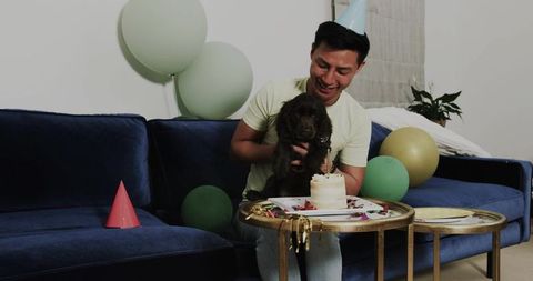 Celebrating man holding puppy and cake on blue sofa with balloons and party hat