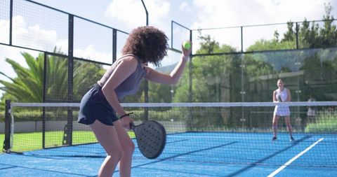 Diverse women playing padel on vibrant blue court tossing ball during outdoor club match