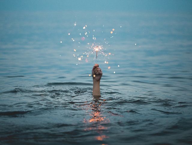 Hand holding sparkler appears from blue ocean, creating surreal and festive scene. Perfect for use in holiday, celebration, and summertime themes. Great for creating a festive mood and highlighting creative and unique concepts in advertisements, websites, and social media content.