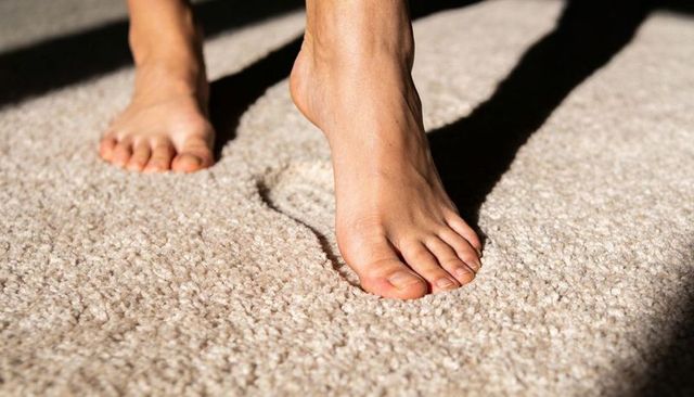 Pressing barefoot feet creating deep indentations in plush beige carpet under sunlight
