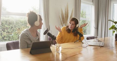 Diverse female co-hosts recording home podcast at sunlit table wearing headphones