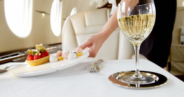 Person arranging elegant fruit tart and meringues on plate beside glass of champagne on white tablecloth in first-class airplane cabin. Perfect for articles on luxury travel, airline amenities, upscale gourmet experiences, and exclusive services.