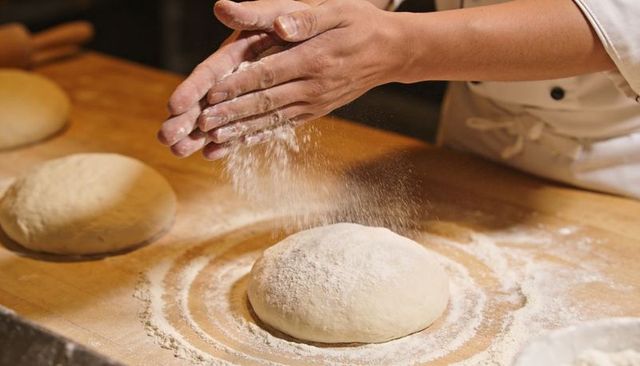 Artisan baker dusting flour on round dough on wooden bench, preparing rustic bread loaves