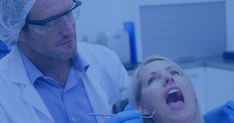 A dentist wearing protective glasses and a hair cap is examining a patient in a dental clinic. The dentist is focused on using dental tools, while the patient is reclined with an open mouth ready for examination. Surrounding elements have a modern and futuristic digital overlay with a blue hue, suggesting technological advancement in medical practices. Great for articles or presentations about dental health essentials, modern healthcare capabilities, or the integration of technology in dentistry.