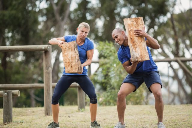 This image depicts two individuals participating in a boot camp obstacle course, carrying heavy wooden logs. Ideal for use in fitness and training promotions, outdoor activity advertisements, or articles on teamwork and physical endurance.