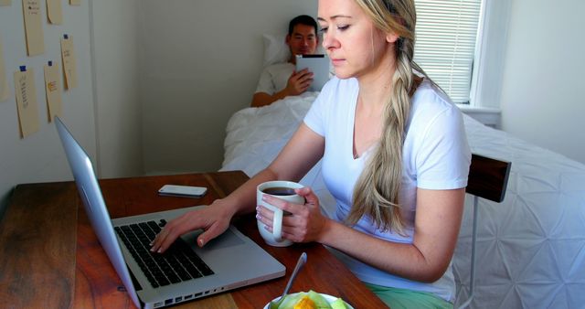 Woman focused on work, typing on laptop, holding coffee cup while a man lounges in bed using his device. Plate with breakfast on table; remote work and productivity themes. Suitable for promotions about work-life balance, modern office setups, or morning routines.