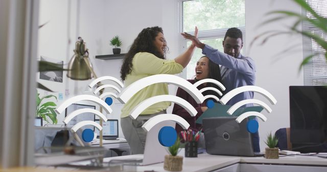 Three professionals in a modern office share a high-five while smiling and surrounded by laptops, indicative of successful teamwork and collaboration. The presence of technology icons overlaid on the image emphasizes the tech-savvy nature of their work. Useful for depicting successful corporate culture, teamwork, and productivity in coworking environments.