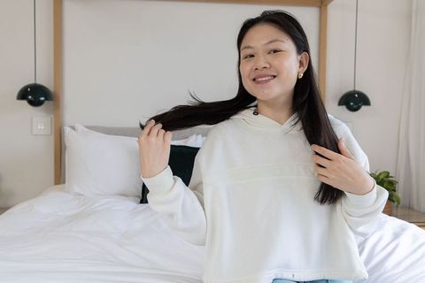 Asian woman smiling styling long hair in minimalist bedroom wearing white hoodie and jeans