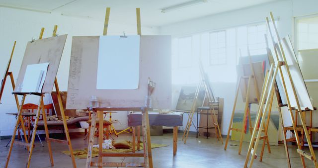 Empty artist studio filled with easels awaiting painters. Sunlight streams through large windows, creating a serene space perfect for creativity. Suitable for illustrating art environments, creative workspaces, artistic inspiration, workshops or hobby rooms.