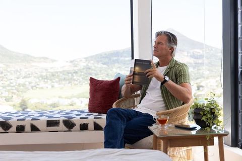 Senior man relaxing with paperback by sunroom window overlooking valley and mountains
