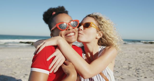 Happy diverse female friends wearing sunglasses kissing and embracing at beach. Vacation, summer, friendship and lifestyle, unaltered.