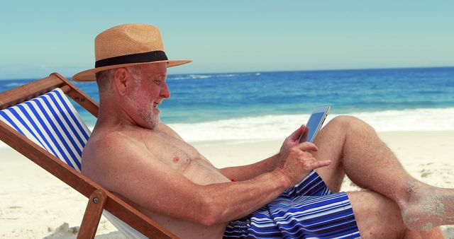 Senior man relaxing on a beach chair by the ocean while using a digital tablet. He is wearing swim shorts and a hat. Ideal for retirement-themed content, travel advertisements, technology usage during vacations, and leisure lifestyle promotions.