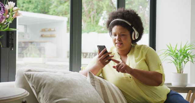 Image of happy plus size african american woman with headphones sitting on sofa with smartphone - Download Free Stock Photos Pikwizard.com