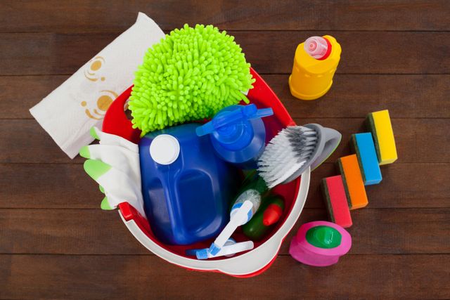 Close-up view of a bucket filled with various cleaning supplies on a wooden floor. Items include sponges, brushes, gloves, detergent bottles, and a microfiber cloth. Ideal for use in articles or advertisements related to household cleaning, sanitation, and hygiene products.