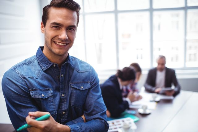This image shows a young businessman in a denim shirt smiling confidently with colleagues in the background engaged in a meeting. Ideal for use in business-related content, articles on teamwork and collaboration, corporate websites, and promotional materials for creative work environments.