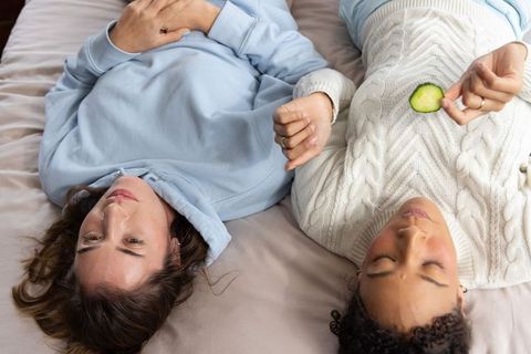 Diverse women lying head-to-head on bed in cozy knitwear holding cucumber slice for skincare