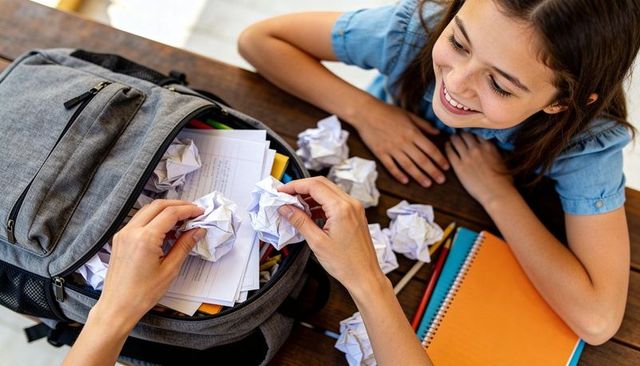 Adult removing crumpled homework from backpack while child watching and smiling