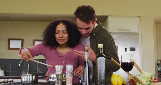 This image captures a joyful and intimate moment of a diverse couple enjoying cooking together in their kitchen. The couple's genuine happiness and affectionate connection are evident as they embrace and smell the delicious meal they are preparing. This visually appealing scene can be used in advertising and promotional materials for cooking products, kitchen appliances, and home lifestyle brands, as well as in editorial content related to relationships, multicultural themes, and domestic life.