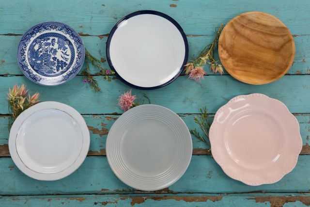 Assorted empty plates arranged on a rustic wooden table with flowers. Ideal for use in home decor, kitchenware, dining, and lifestyle blogs or websites. Perfect for illustrating articles on table settings, vintage kitchenware, or rustic home decor ideas.