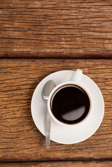 This image shows an overhead view of a cup of coffee on a wooden table, with a spoon resting on the saucer. Ideal for use in articles or advertisements related to coffee, morning routines, relaxation, or rustic settings. Perfect for blogs, social media posts, or websites focused on beverages and lifestyle.