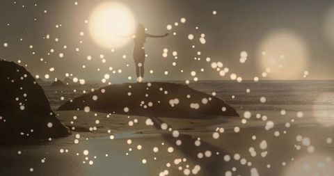 Standing silhouette facing glowing sun over rocky shore, wet sand, waves and bokeh lights