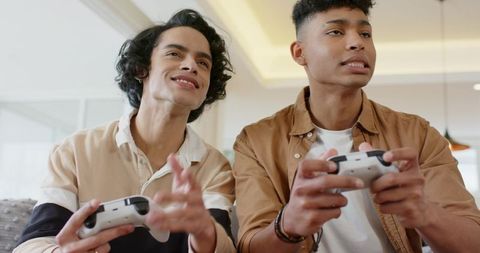 Diverse young men playing video games on couch with white controllers in cozy living room