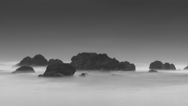 Monochrome image showing large rocks emerging from misty water at dawn, creating a serene and tranquil seascape. Suitable for use in interior decoration, inspirational backgrounds, and mindfulness content.