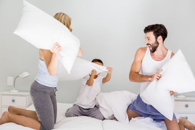 Family enjoying a playful pillow fight on bed in bedroom. Parents and child laughing and having fun. Perfect for themes related to family bonding, indoor activities, happiness, and casual home life.