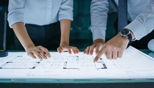 Architects collaborating over illuminated floor plan on drafting table, hands pointing, teamwork